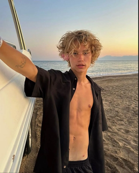 A shirtless young man standing next to a car on the beach.