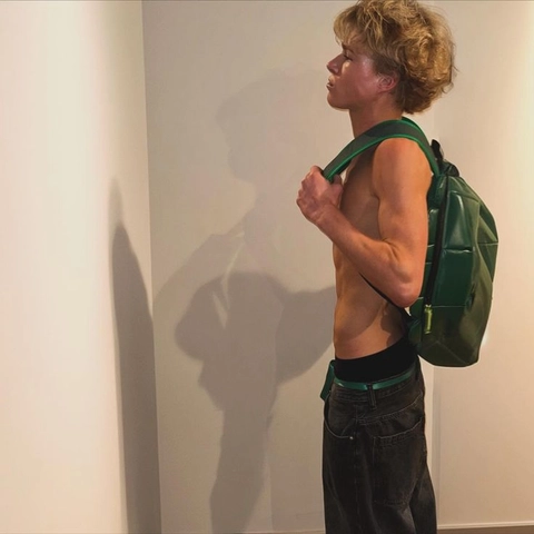 A shirtless man with a backpack standing in front of a wall.