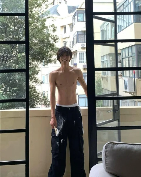 A shirtless man standing in front of a window.