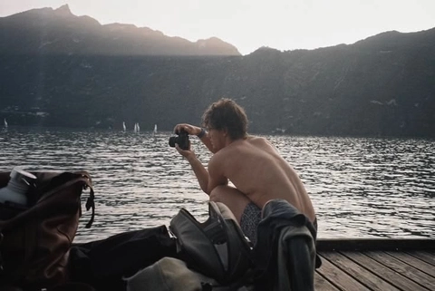 A shirtless man sitting on a dock taking a picture of himself.