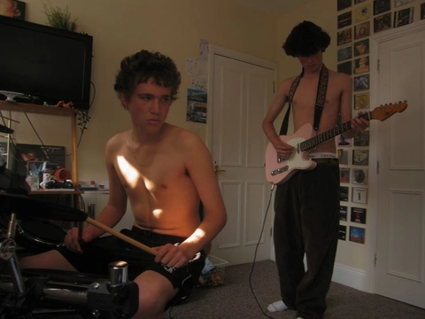 A shirtless young man playing a guitar while another man plays a guitar.