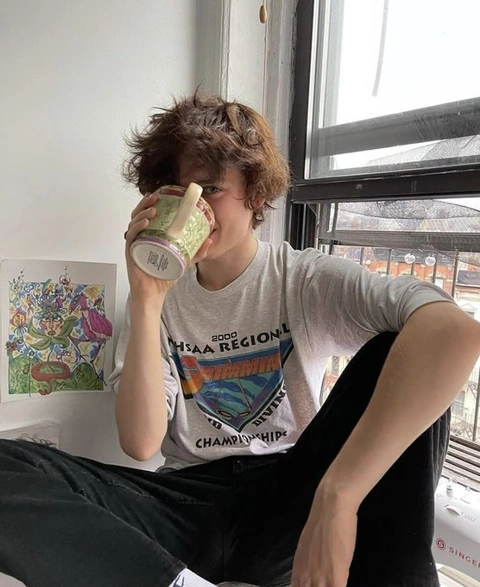 A young man sitting on a window sill drinking from a can.