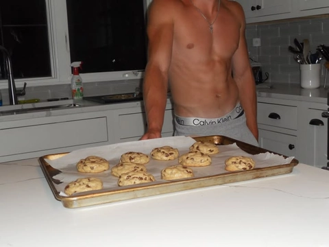 A shirtless man standing in a kitchen holding a tray of cookies.