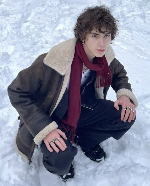 A young man sitting in the snow wearing a brown jacket and scarf.